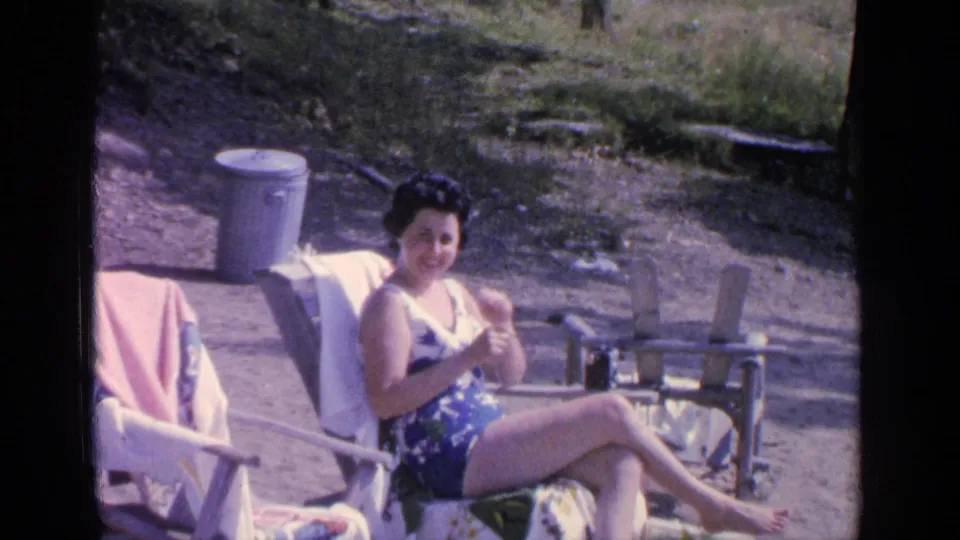 At Greenwood Lake in 1964, a woman smiles from a wooden beach chair on a sunny lakeside beach with towels and another chair nearby.