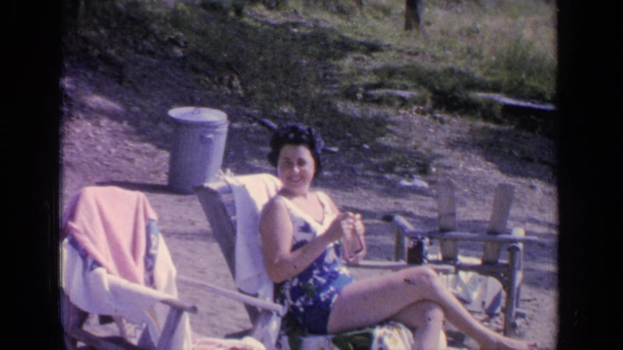 Woman in a blue patterned swimsuit seated in a chair outdoors beside another towel-draped chair.