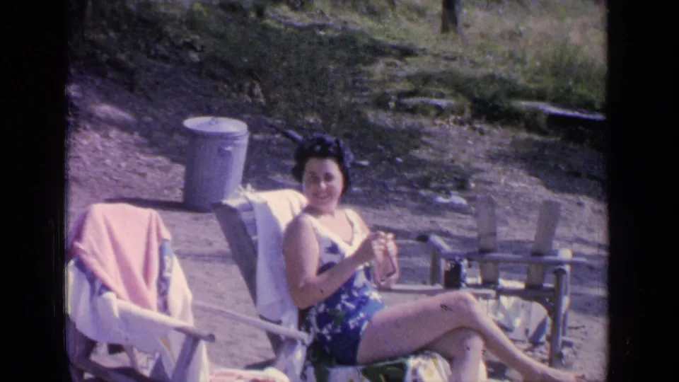 At Greenwood Lake in 1964, a woman in a blue patterned swimsuit sits in a lawn chair near a pink towel and metal trash can.