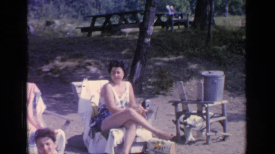 At Greenwood Lake in 1964, the woman sits with legs crossed in a chair, with picnic tables and trees behind her.
