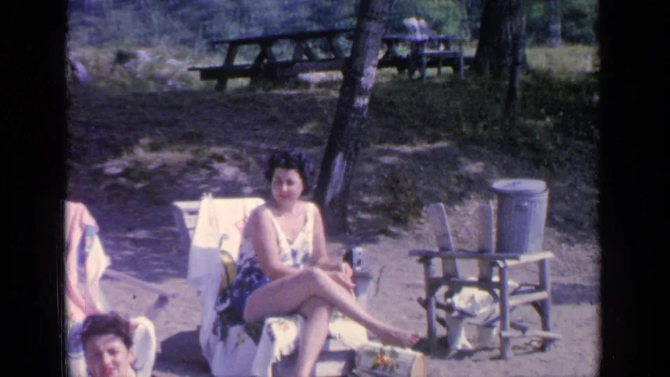 At Greenwood Lake in 1964, the woman remains seated outdoors near a metal trash can, wooden chairs, trees, and picnic tables.