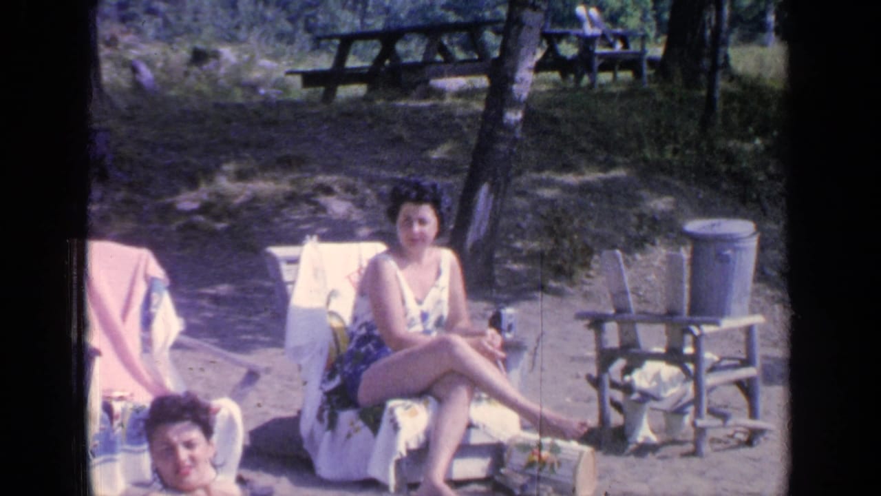 Two adult women relax on beach chairs in a wooded lakeside area.