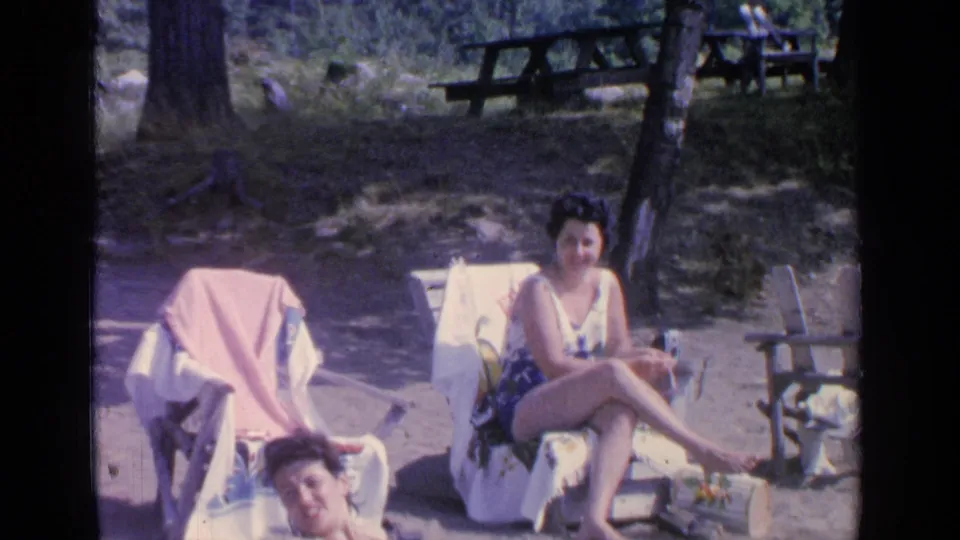 1964 Greenwood Lake home-movie frame of two adult women relaxing beside towel-covered chairs in a shaded outdoor beach area.