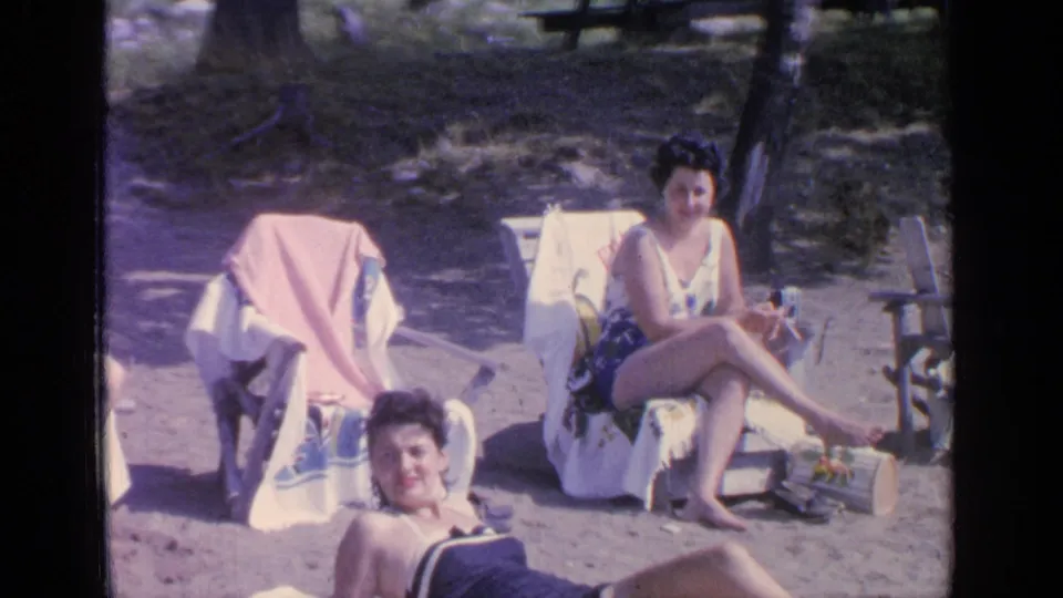1964 Greenwood Lake frame showing a reclining adult woman in the foreground and a seated woman behind her near beach chairs.
