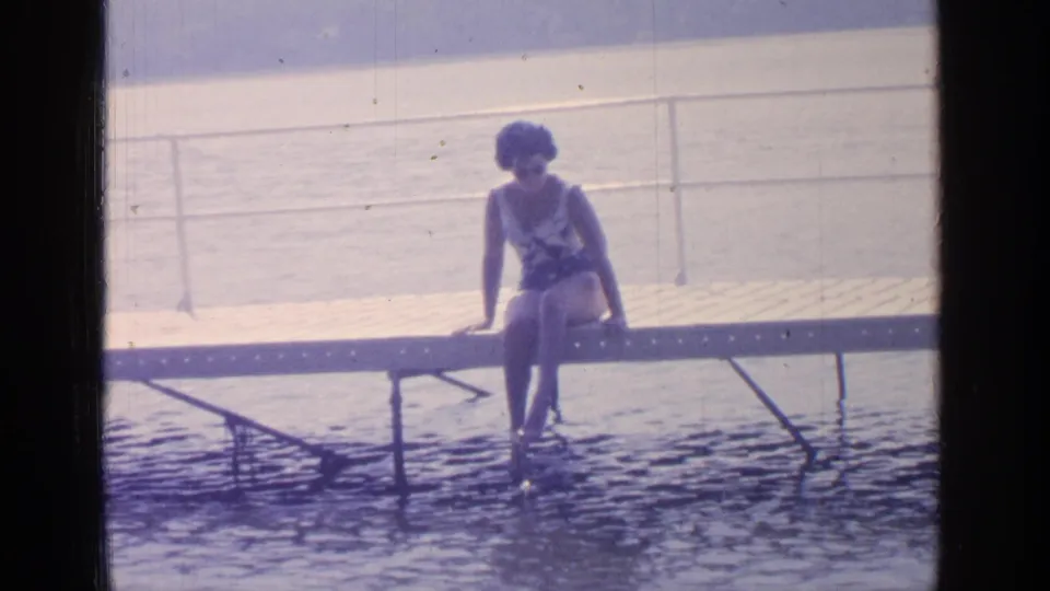 In 1964 Greenwood Lake footage, a woman in a one-piece swimsuit sits on a wooden dock above rippling water.