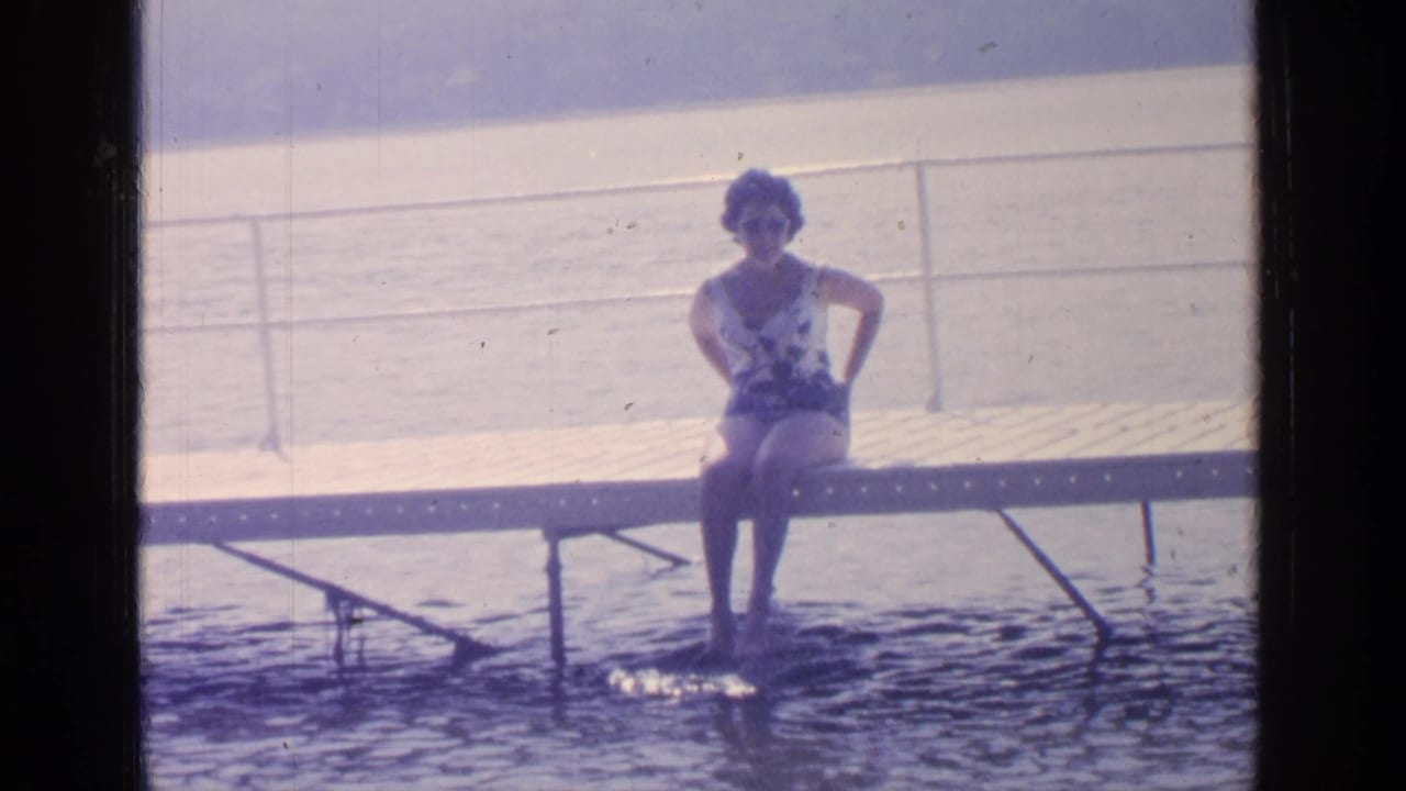 Woman seated on the dock, facing the camera with lake and railings behind her.