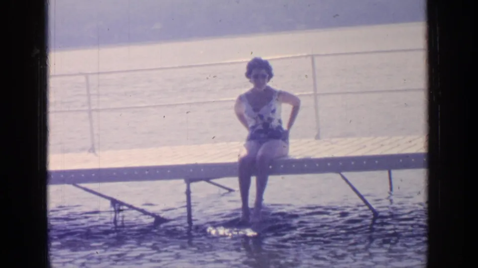 A woman sits on a Greenwood Lake dock in 1964, facing the camera with her feet in the water and hills beyond.