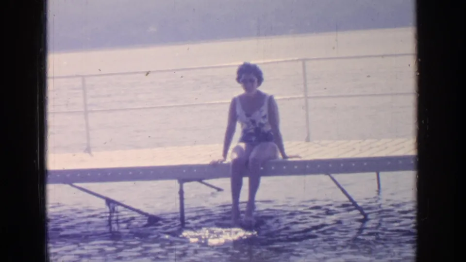 The 8mm frame shows the woman seated upright on the Greenwood Lake dock, legs lowered toward the water.