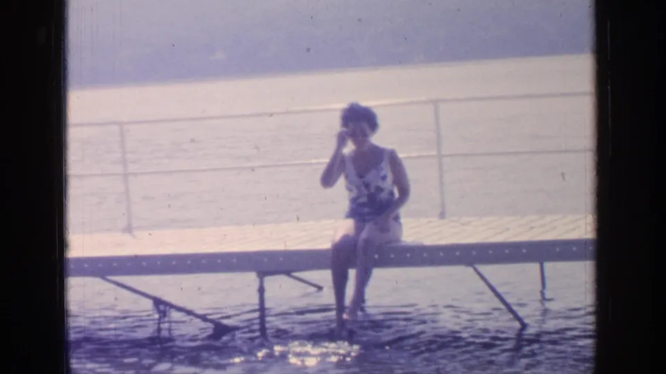 In the final still, the woman remains seated on the Greenwood Lake dock and lifts a hand near her face as water glints below.
