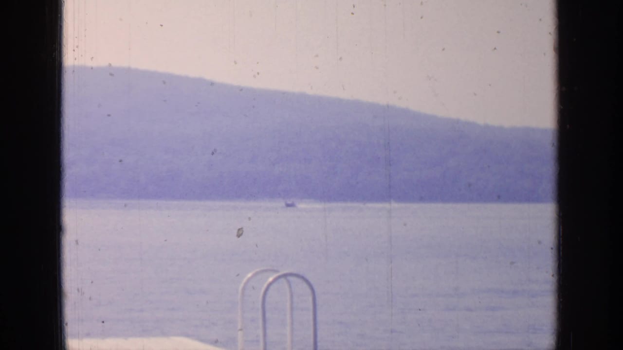 Greenwood Lake, New York, appears beyond dark window framing, with a distant small boat, pale sky, and hazy mountain shoreline.