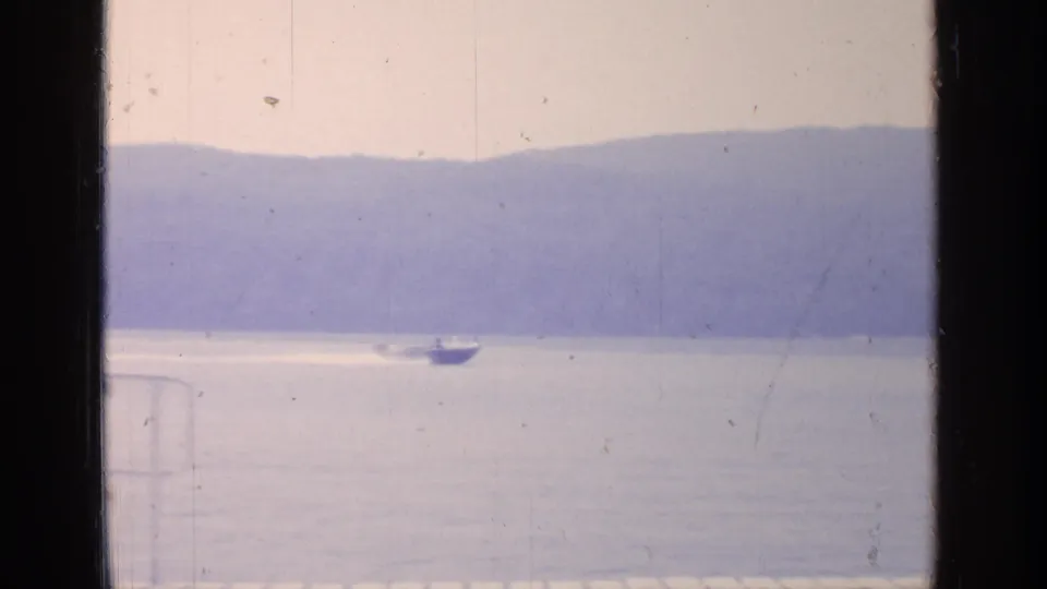 A small boat leaves a bright wake across Greenwood Lake, with hazy wooded hills behind it and a pool ladder partly visible in the foreground.