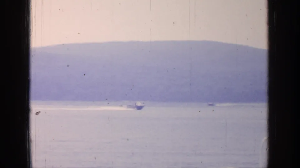 A small boat travels across Greenwood Lake near the far shoreline, with a bright wake, hazy mountains, and dark window framing in the scan.
