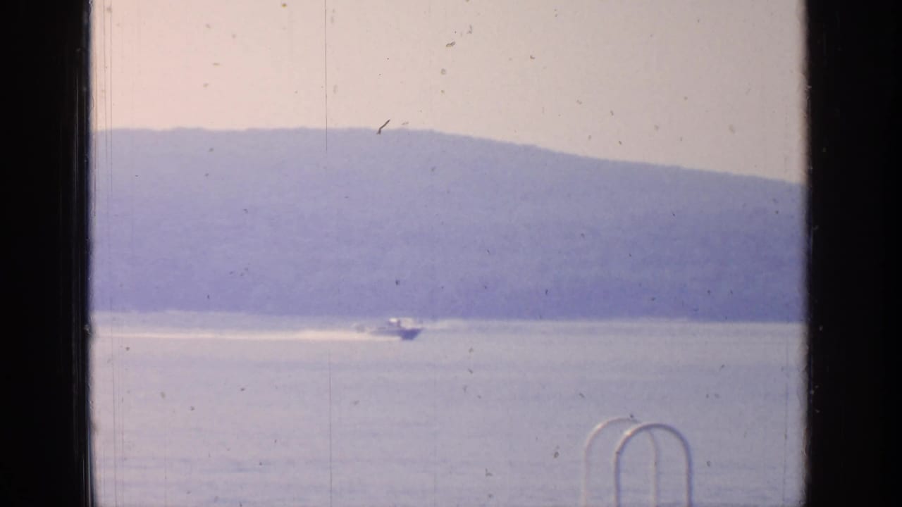 1964 Greenwood Lake home-movie frame showing a small boat moving across calm water beneath hazy hills, with a dock ladder in front.