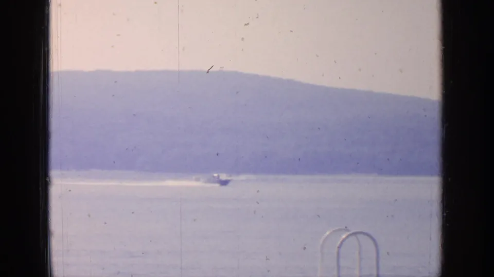 1964 Greenwood Lake home-movie frame showing a small boat moving across calm water beneath hazy hills, with a dock ladder in front.