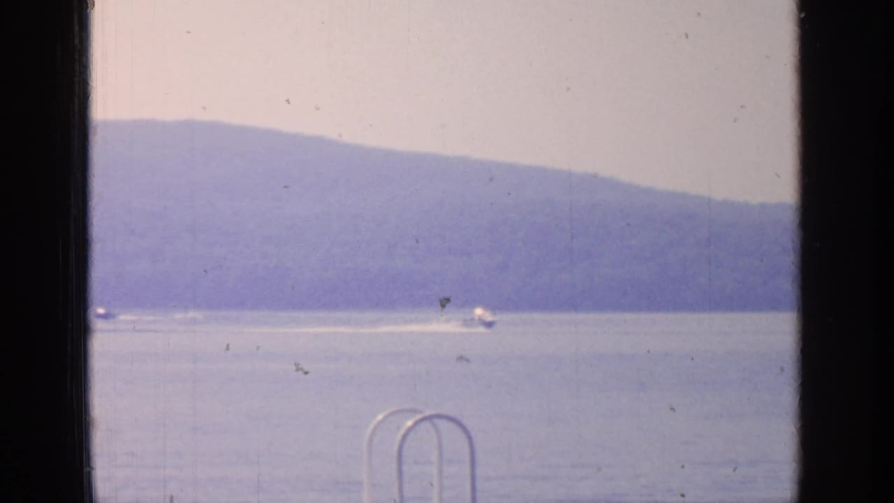 1964 Greenwood Lake frame with small boats crossing the water against a hazy hillside backdrop and dock hardware at the edge.
