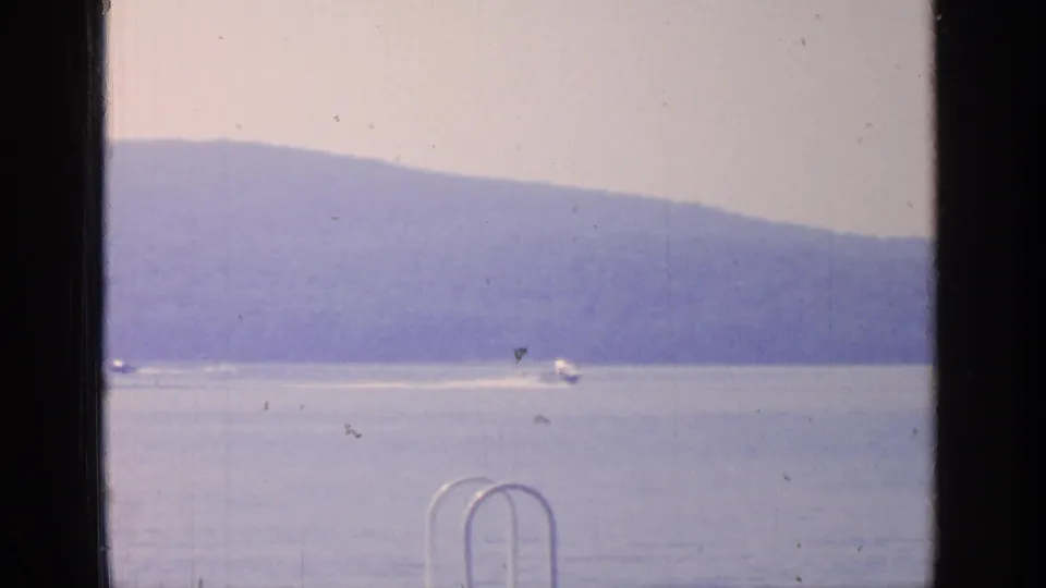 1964 Greenwood Lake frame with small boats crossing the water against a hazy hillside backdrop and dock hardware at the edge.