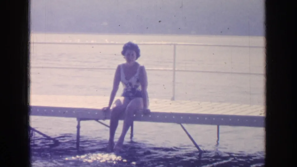 1964 Greenwood Lake frame showing a young woman seated on a wooden dock in a patterned one-piece swimsuit, feet in the water.