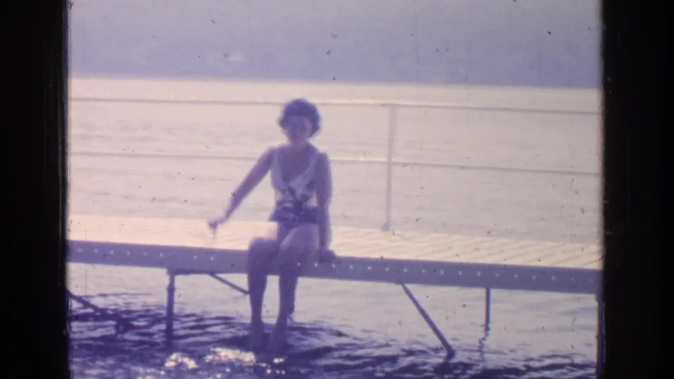 1964 Greenwood Lake home-movie frame of a young woman on a dock waving toward the camera, with railings and lake behind her.