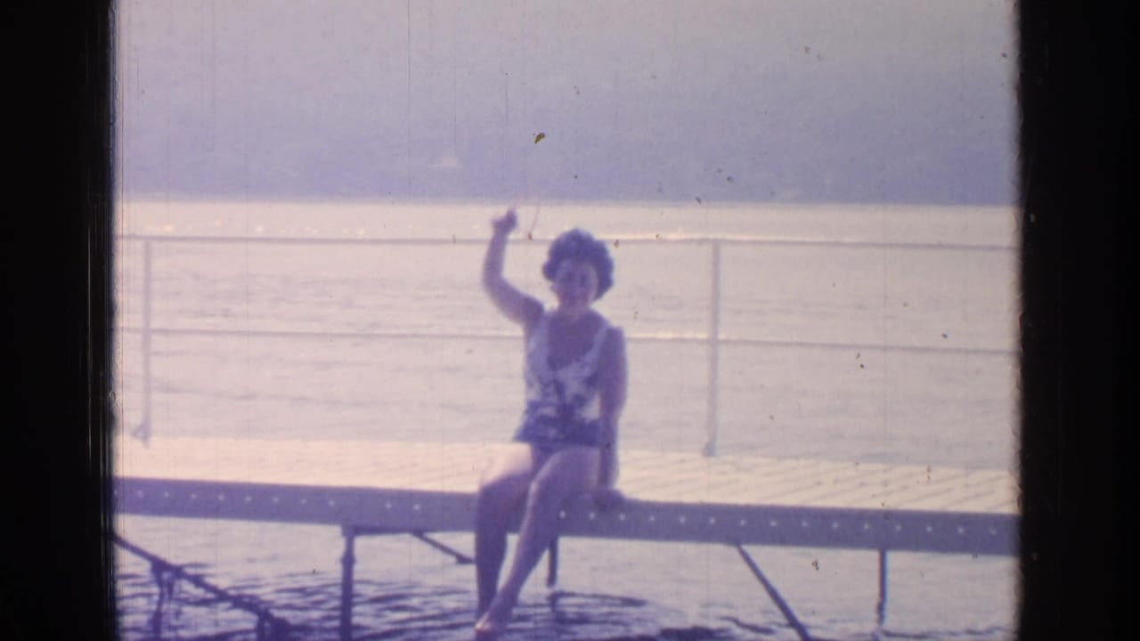 1964 Greenwood Lake home-movie frame showing a woman in a patterned swimsuit seated on a dock, with her feet near the water and one hand raised.