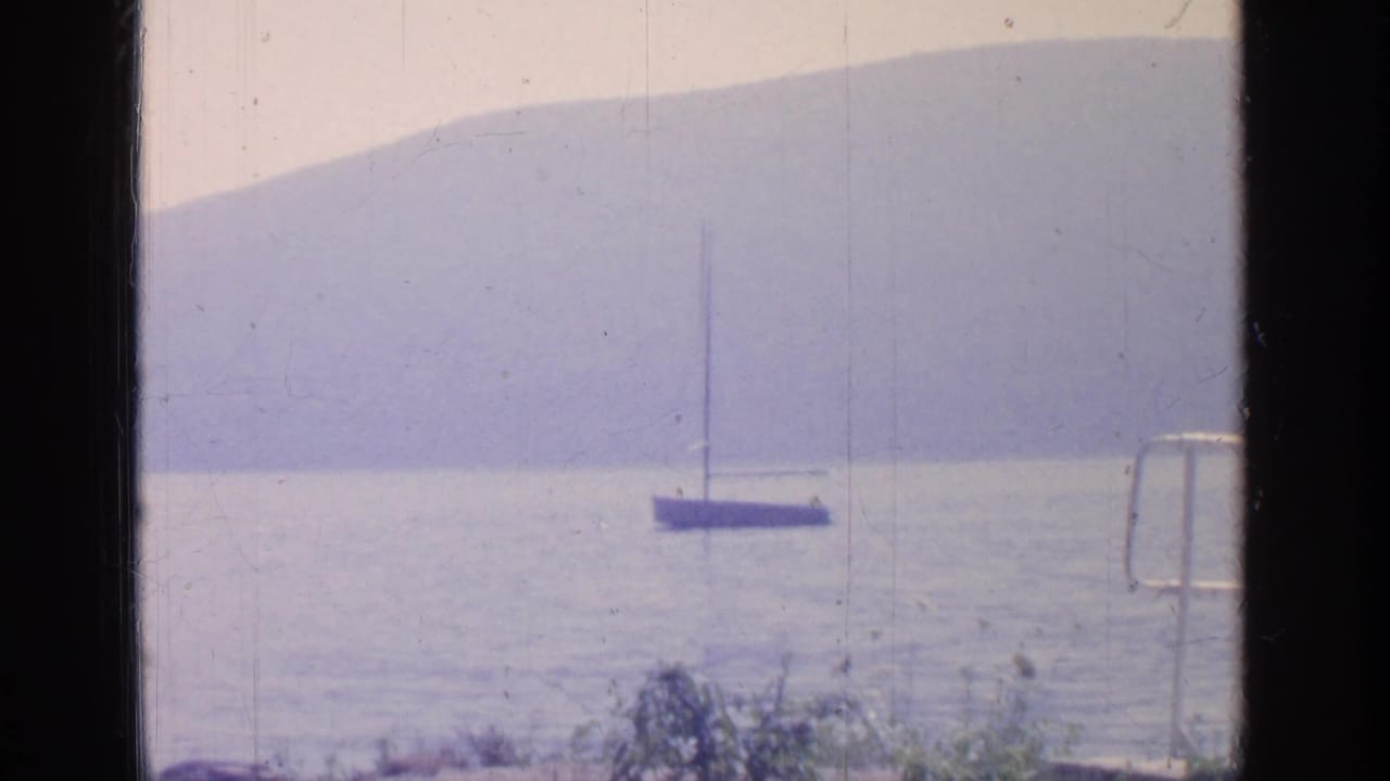 Greenwood Lake view from the 1964 home movie, with a small sailboat on calm water, lakeside railing, and hazy hills beyond.