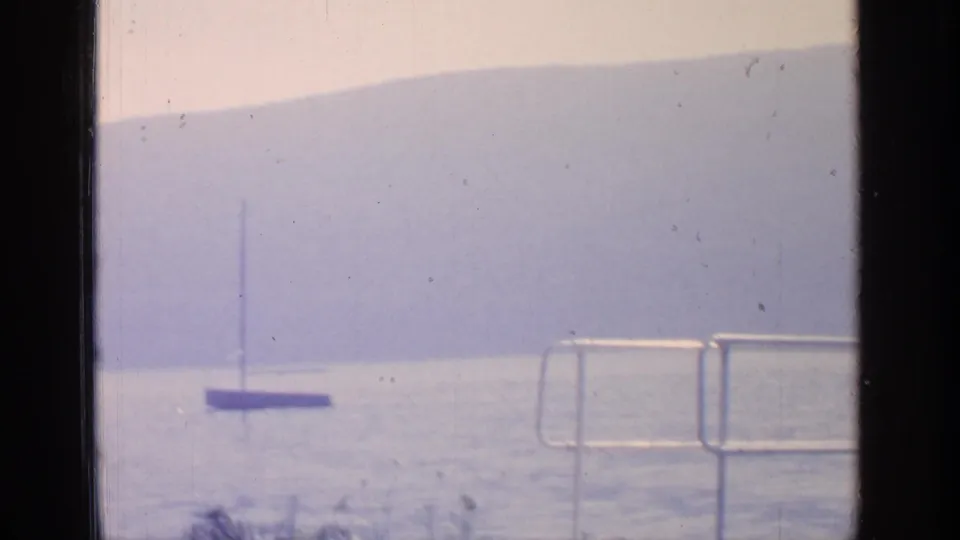 1964 Greenwood Lake frame showing a small sailboat near a waterfront railing, with calm water and hazy hills across the lake.
