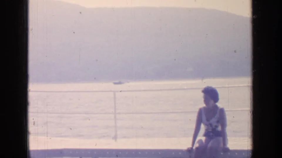 At Greenwood Lake in 1964, a woman in white swimwear sits on a metal bench beside the waterfront railing.