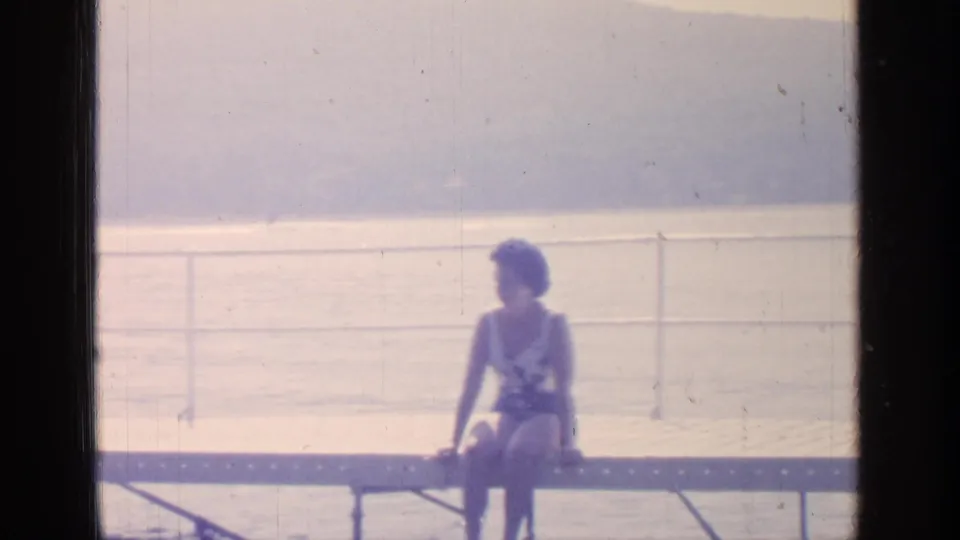 A 1964 Greenwood Lake home-movie frame shows a woman seated on a metal bench in white swimwear, facing the waterfront.