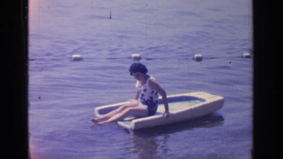 Adult woman in a floral swimsuit sits on a small float in Greenwood Lake water, with white buoys marking the swim area.