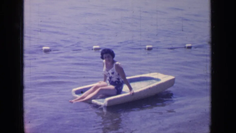 In this 1964 Greenwood Lake frame, a woman sits upright on a pale float inside a buoy-marked swimming area.