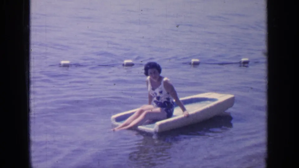 A woman in a floral swimsuit sits low on a small float at Greenwood Lake, framed by calm water and a buoy rope.