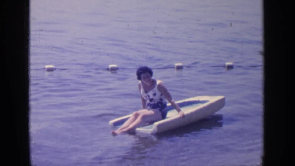 The adult woman remains seated on the float in Greenwood Lake, with buoys and rope visible across the background water.
