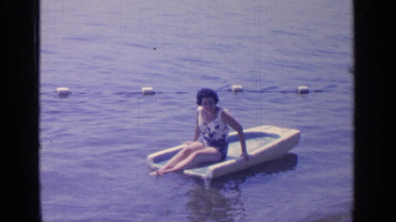 At Greenwood Lake in 1964, a woman in a floral swimsuit sits on a small white and blue raft near a buoy line.