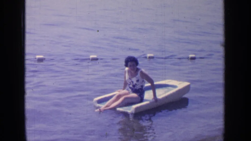 At Greenwood Lake in 1964, a woman in a floral swimsuit sits on a small white and blue raft near a buoy line.