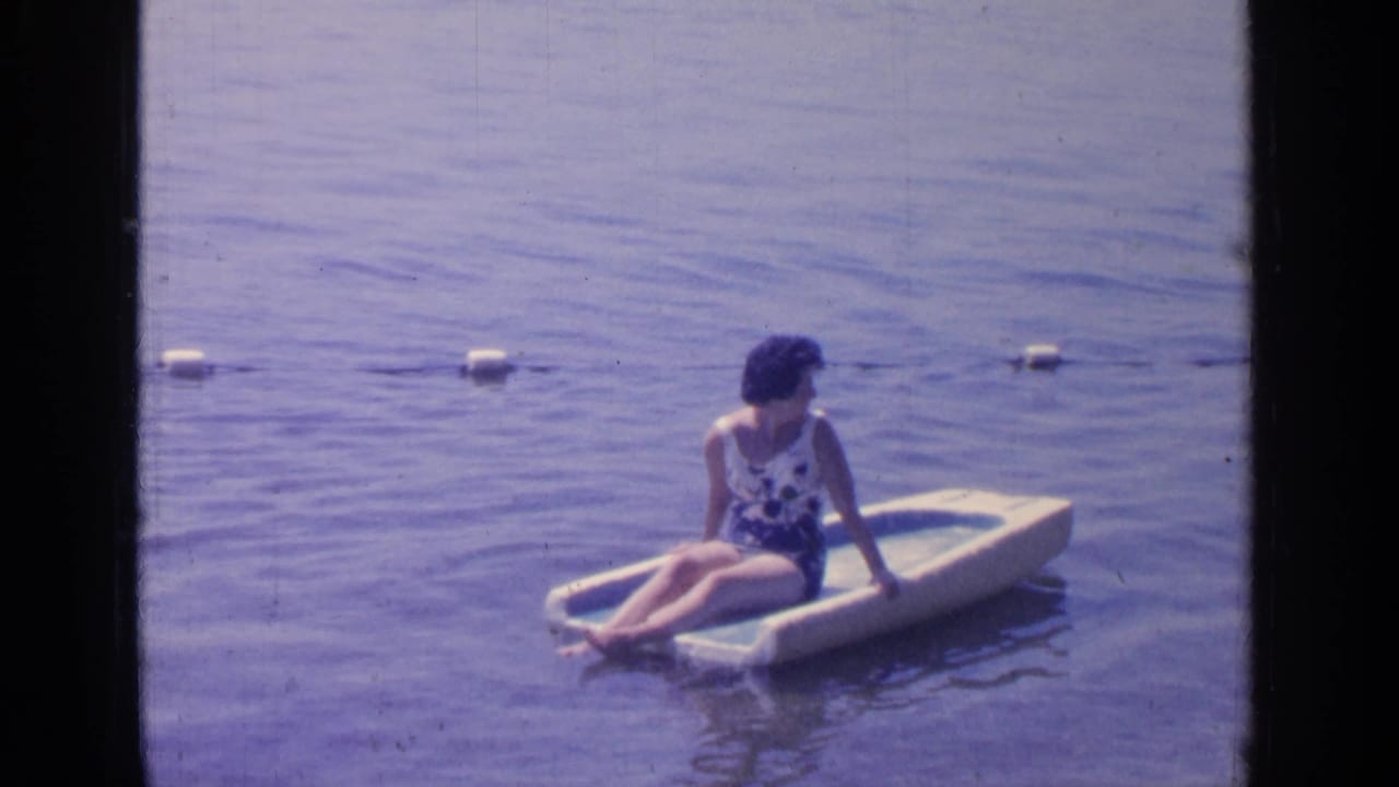 The woman remains seated on the raft at Greenwood Lake, with a line of white buoys stretching across the background.
