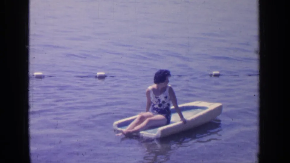 The woman remains seated on the raft at Greenwood Lake, with a line of white buoys stretching across the background.