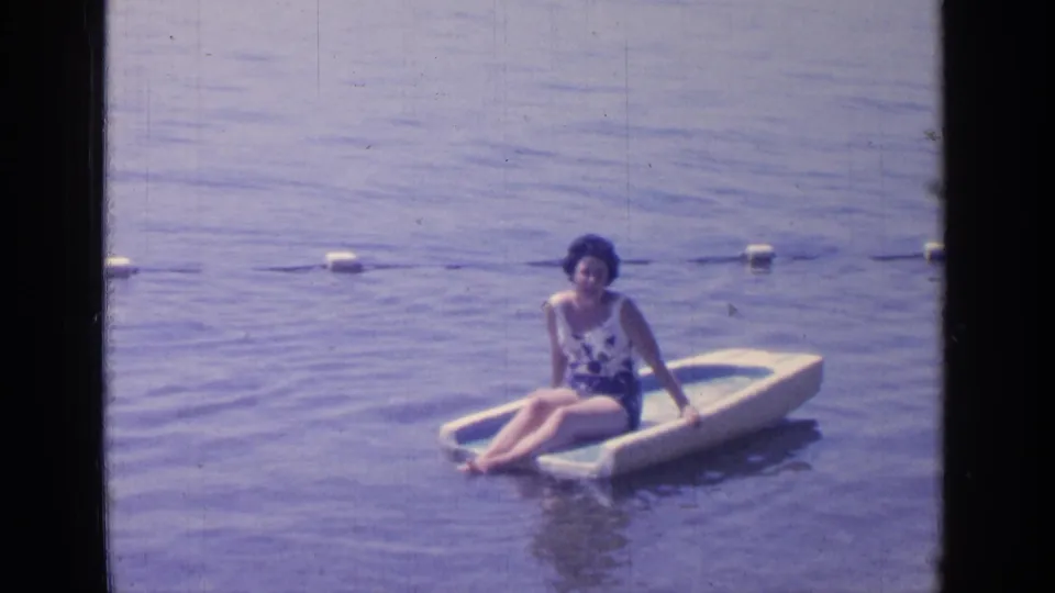 A 1964 8mm frame shows the woman facing the camera while seated on the raft, with lake water and buoys behind her.