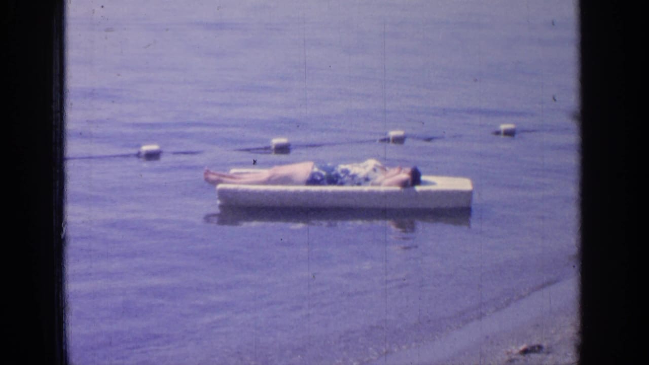 1964 Greenwood Lake home-movie frame showing a man reclining on a white floating platform beside a buoy line.
