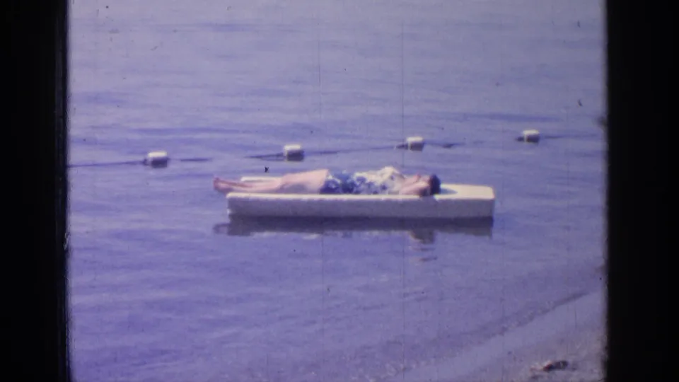 1964 Greenwood Lake home-movie frame showing a man reclining on a white floating platform beside a buoy line.