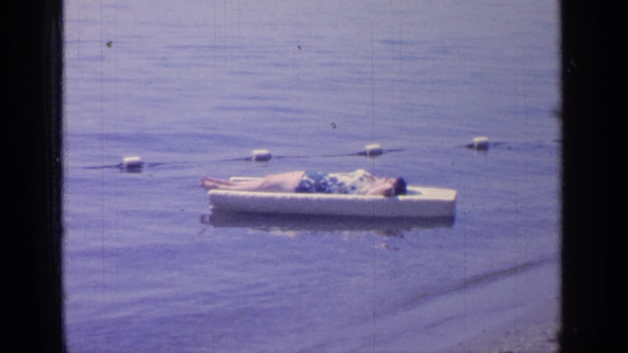 Man sunbathing on a white platform floating in a lake with buoys behind him.