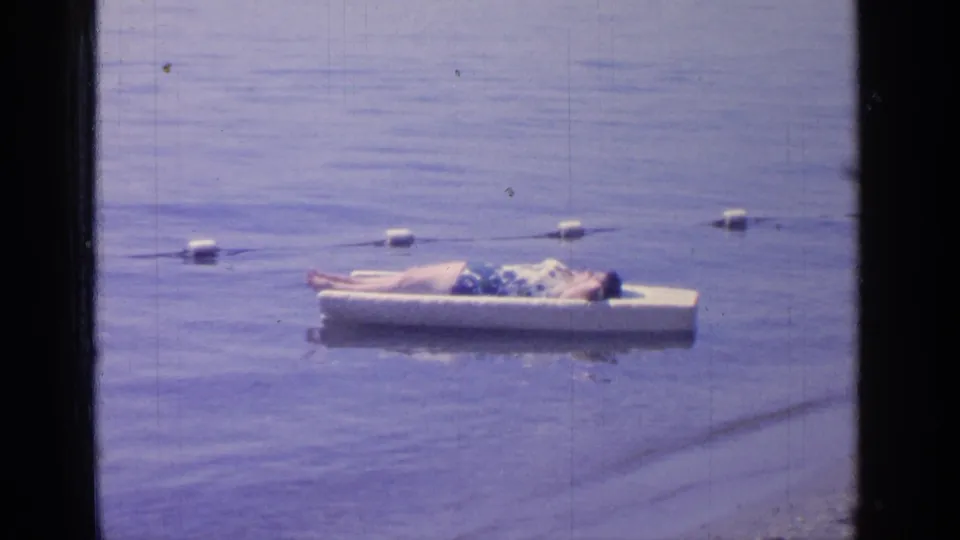 Home-movie frame from Greenwood Lake, New York, showing a man lying back on a white float in 1964.