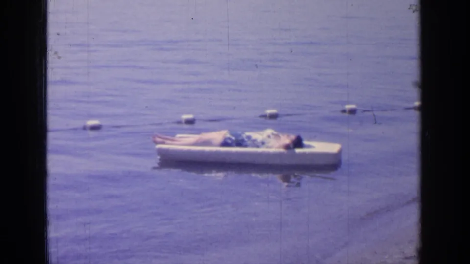 A 1964 8mm frame of a man resting on a white floating platform on Greenwood Lake near a row of buoys.