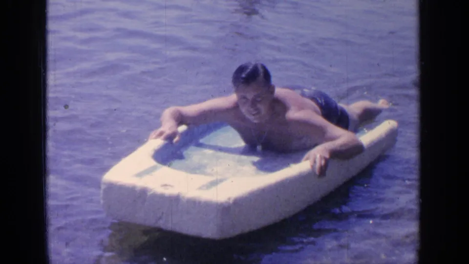 At Greenwood Lake in 1964, an adult man lies on a white foam float while holding its sides in the water.