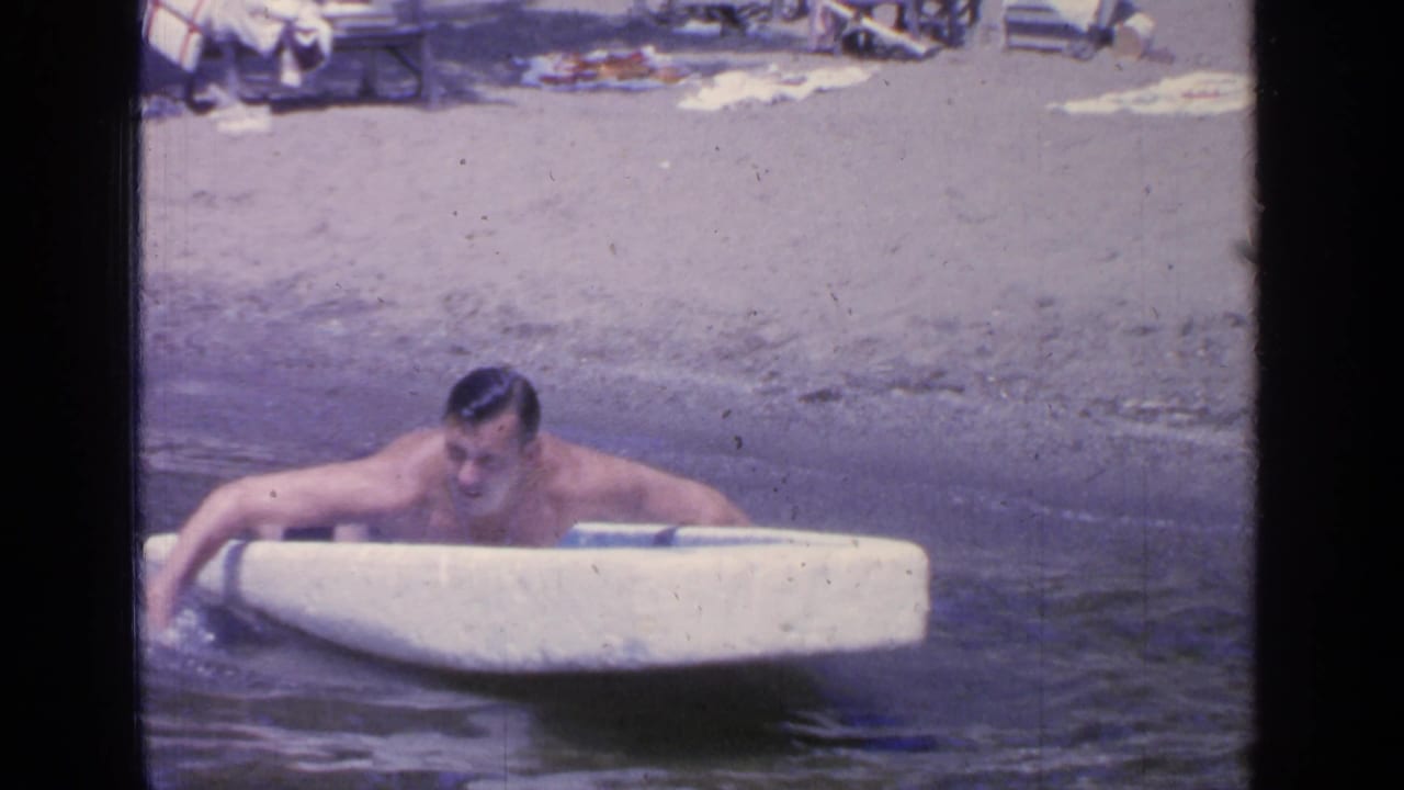 At Greenwood Lake in 1964, a man leans over a small white boat while floating near a sandy beach with lounge chairs behind him.