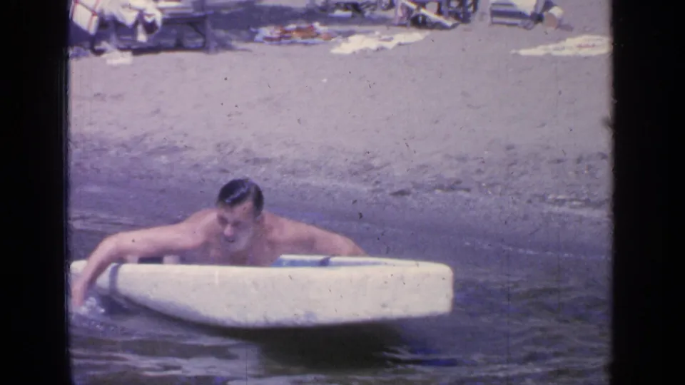 At Greenwood Lake in 1964, a man leans over a small white boat while floating near a sandy beach with lounge chairs behind him.
