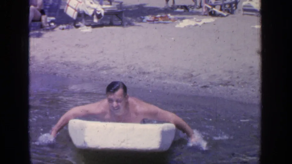Facing the camera, the man continues moving the small white boat by hand near the Greenwood Lake shoreline in this 1964 home-movie frame.
