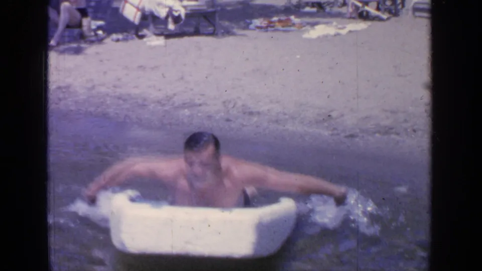 A softer 8mm frame shows the man splashing forward in the small white boat, with Greenwood Lake beach details faintly behind him.