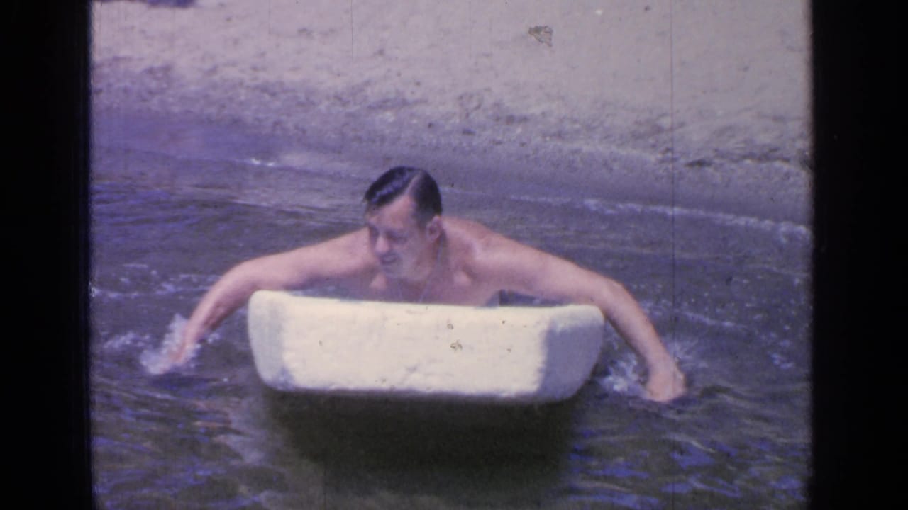 At Greenwood Lake in 1964, a shirtless adult man paddles in shallow water while gripping a white foam board.