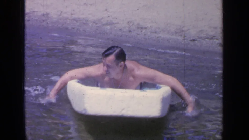 The adult swimmer continues across shallow Greenwood Lake water with both arms around a white foam board.