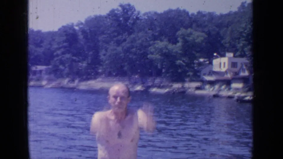 A shirtless adult man faces the camera in Greenwood Lake water, with trees and lakeside houses visible behind him.