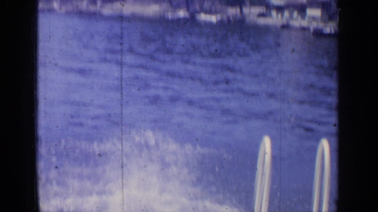 1964 Greenwood Lake home-movie frame showing blue water, a white dock ladder, and faint shoreline detail.